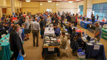 In a crowded event hall, participants attend a career fair. There are rows of tables with employers.