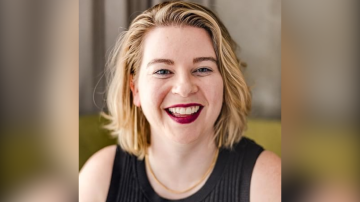 Tori Dunlap, an internationally recognized money and career expert and podcast host, has short blonde hair and red lipstick. She smiles at the camera. She is wearing a black top and a gold necklace. The background is blurred.