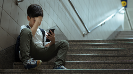 Asian teenage boy sitting at stair at school, covering his face with hands, face down, holding smartphone in low light feeling frustrated, lonely, stress and depressed. Cyber bullying concept.
