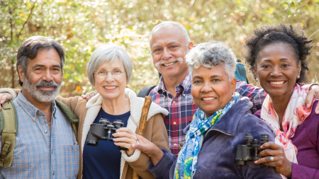 Active senior adult friends hiking in wooded forest area. The multi-ethnic group wears a backpack and uses binoculars to explore nature around them. Middle Eastern, Latin, African and Caucasian ethnicities. 60s to 70s age range.