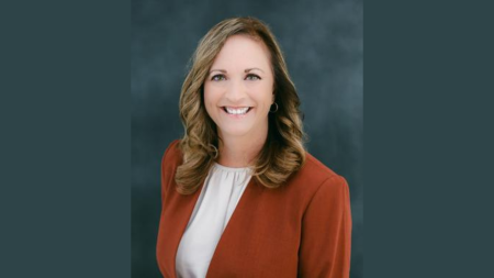 Dr. Veronica Kelley taking a portrait photo in front of a dark blue and dark grey mixed gradient background.  Dr. Veronica Kelley smiling, wearing a white blouse with a burnt orange blazer.