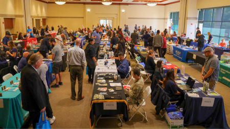 In a crowded event hall, participants attend a career fair. There are rows of tables with employers.