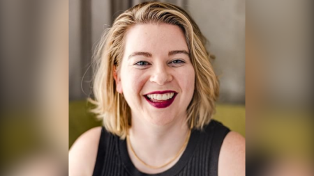 Tori Dunlap, an internationally recognized money and career expert and podcast host, has short blonde hair and red lipstick. She smiles at the camera. She is wearing a black top and a gold necklace. The background is blurred.